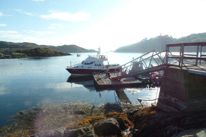 Boat docked at a pier with calm water and hills in the background on a sunny day.