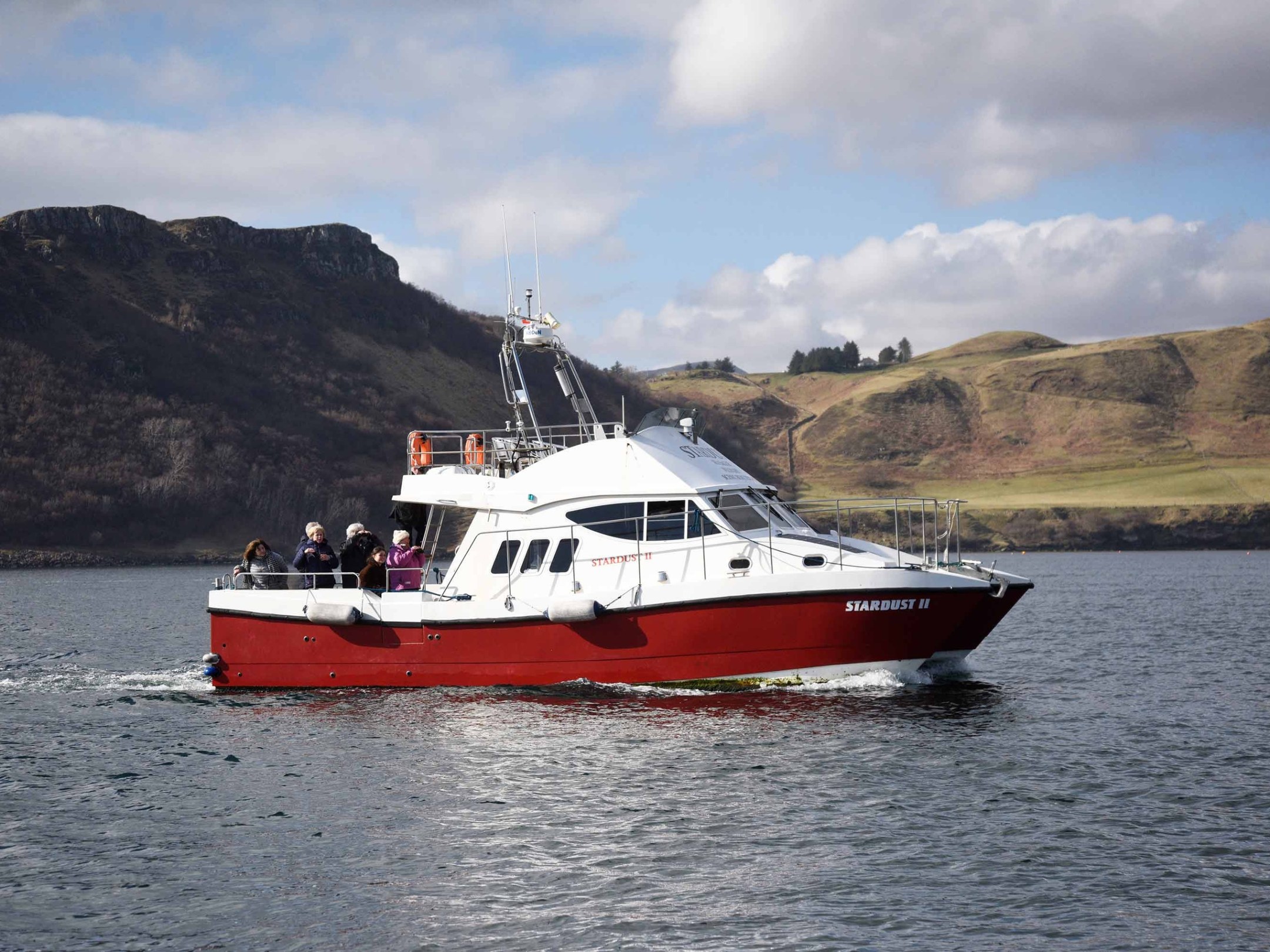 Red and white boat with passengers on calm water, hilly landscape in background.
