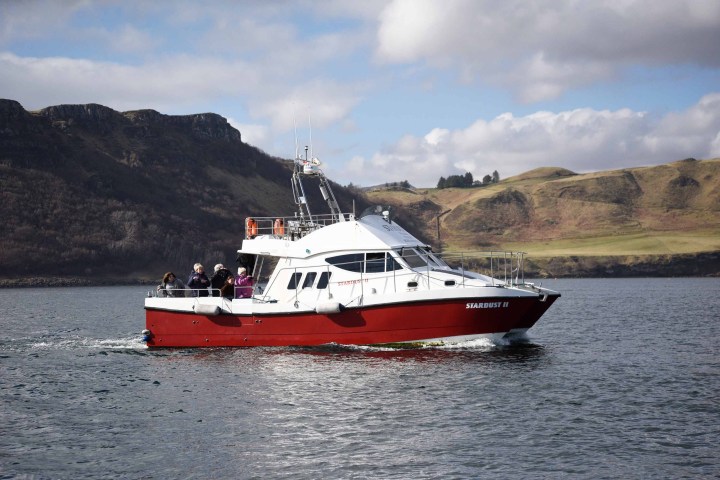 Red and white boat with passengers on calm water, hilly landscape in background.