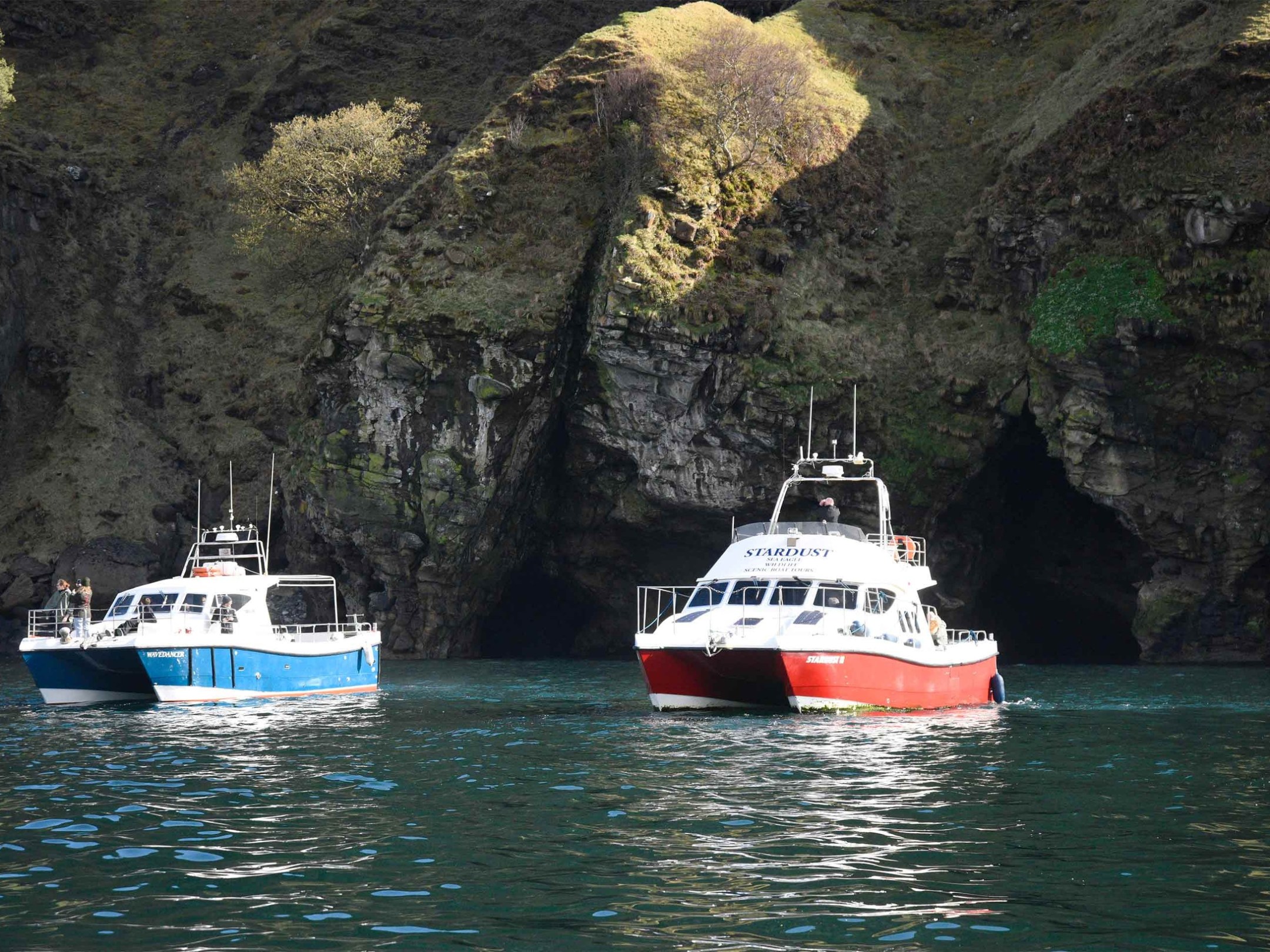 Two boats floating near a rocky coastline with sea caves in the background.