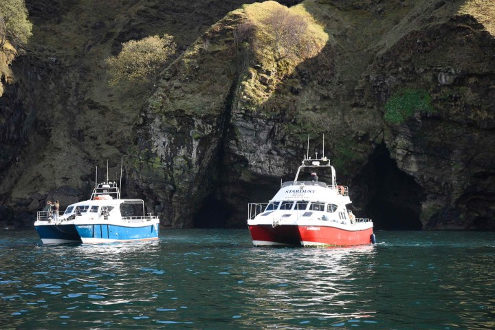 Two boats floating near a rocky coastline with sea caves in the background.