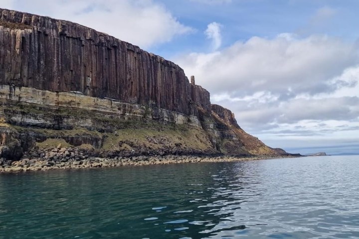 Steep rocky cliffside with vertical striations next to calm blue water under a partly cloudy sky.