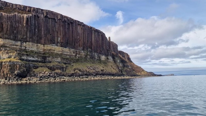 Steep rocky cliffside with vertical striations next to calm blue water under a partly cloudy sky.