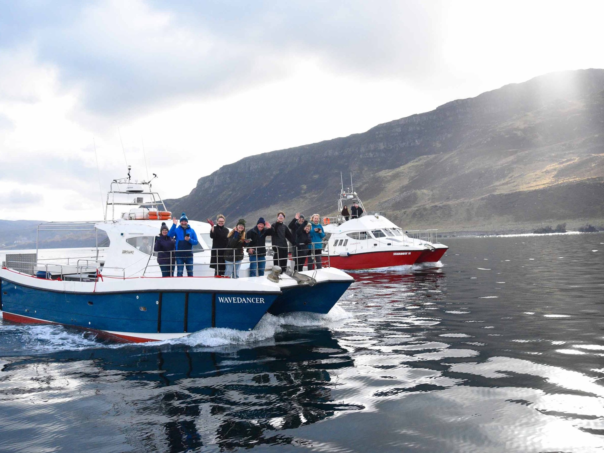 Two boats with people on a lake, mountain in background, cloudy sky.