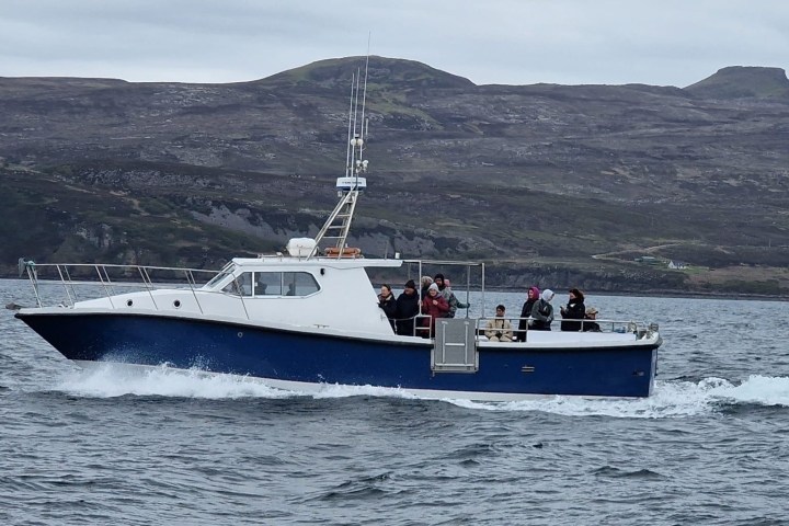A blue and white boat with people onboard is moving across a body of water near a mountainous coastline.
