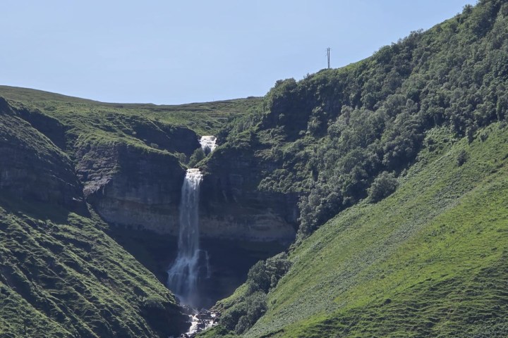 Waterfall flowing between lush green hills under a clear blue sky.